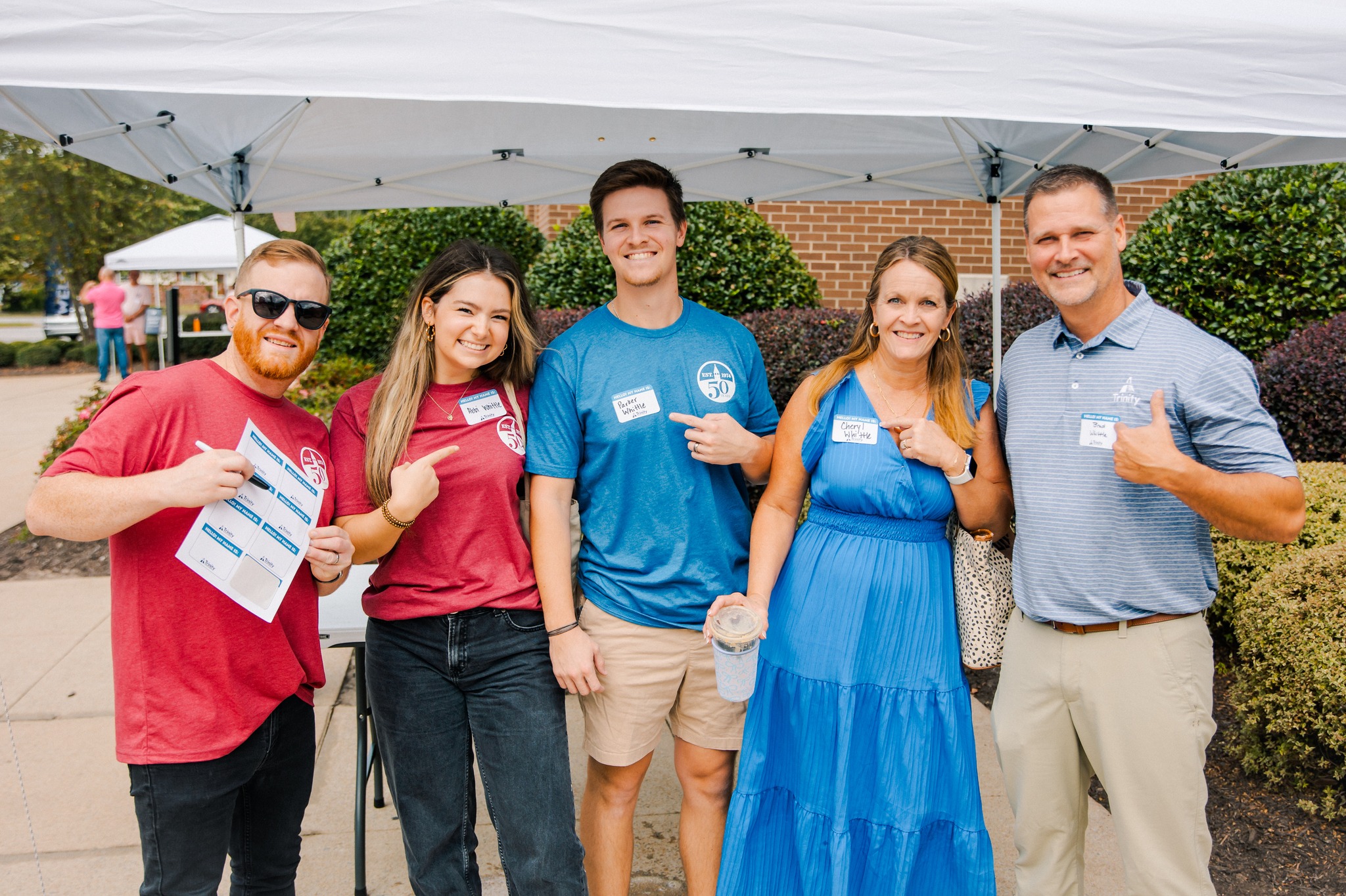 A group of five smiling people wearing name tags gathered under a tent outside Trinity Baptist Church at a church event