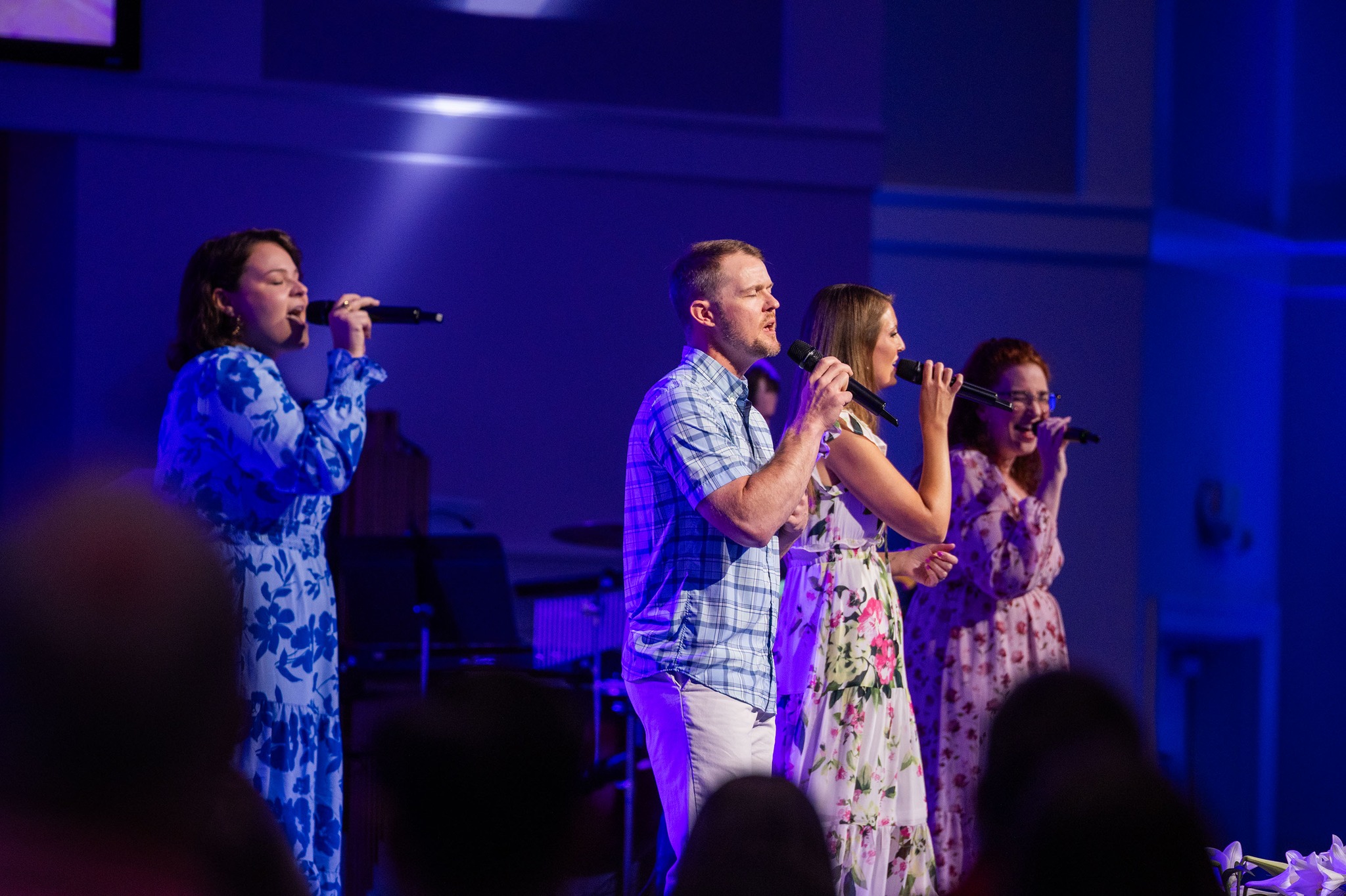 Four worship team members singing on stage with microphones under blue stage lighting at Trinity Baptist Church